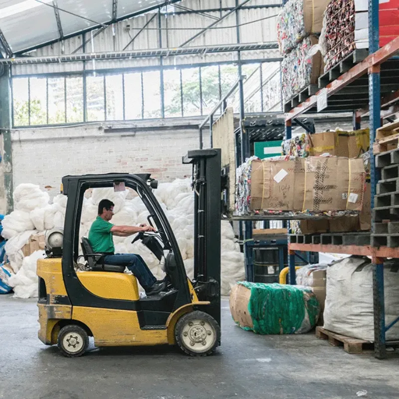 Worker operating a yellow forklift near stacked boxes and baled recycling material in a warehouse