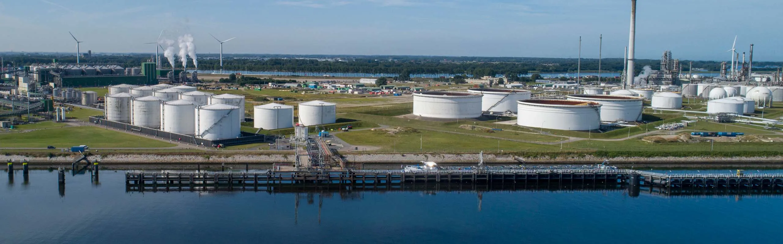Panoramic view of an industrial tank farm along a waterway with smoke stacks in the distance
